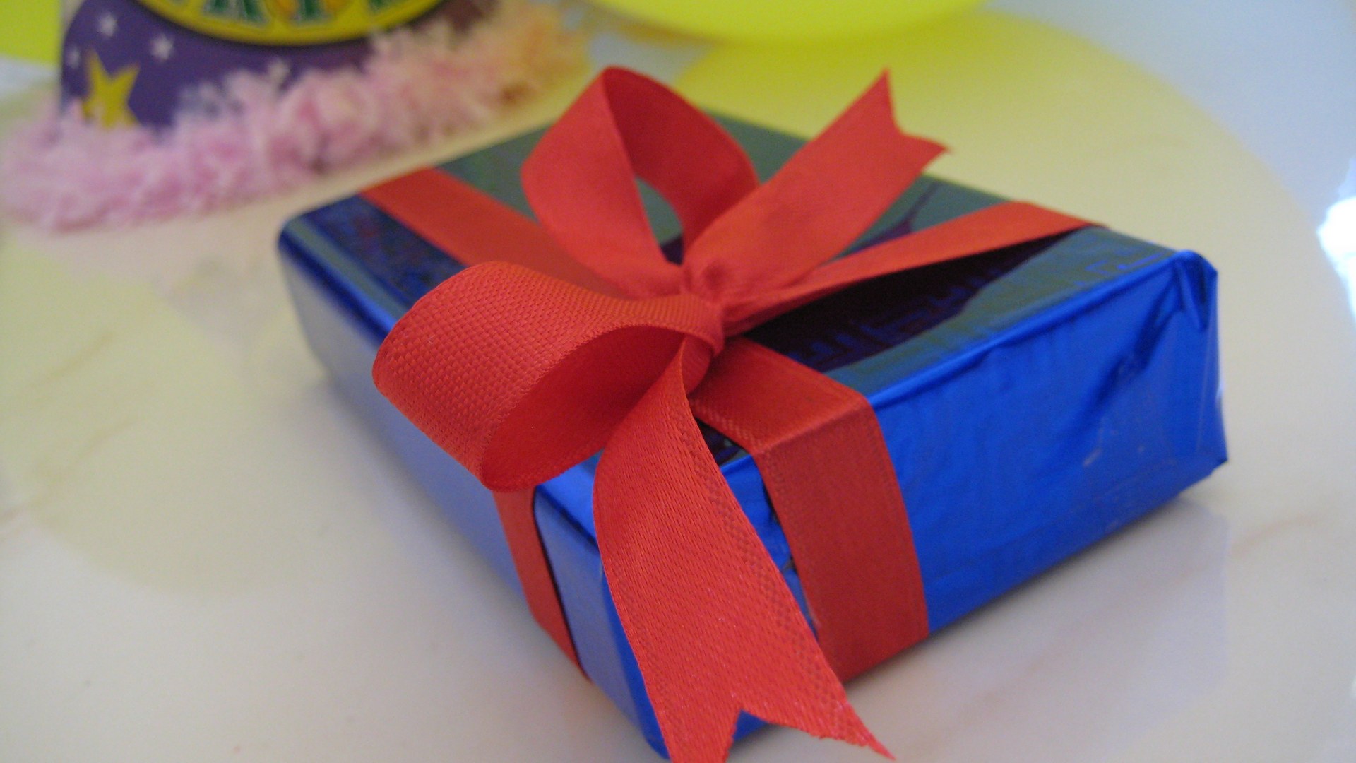 Regalo envuelto en papel azul brillante con un lazo rojo, junto a un gorro de cumpleaños y un globo amarillo